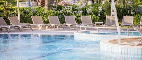 Zona de piscina con sillas de descanso y sombrillas, rodeada de flores y palmeras.