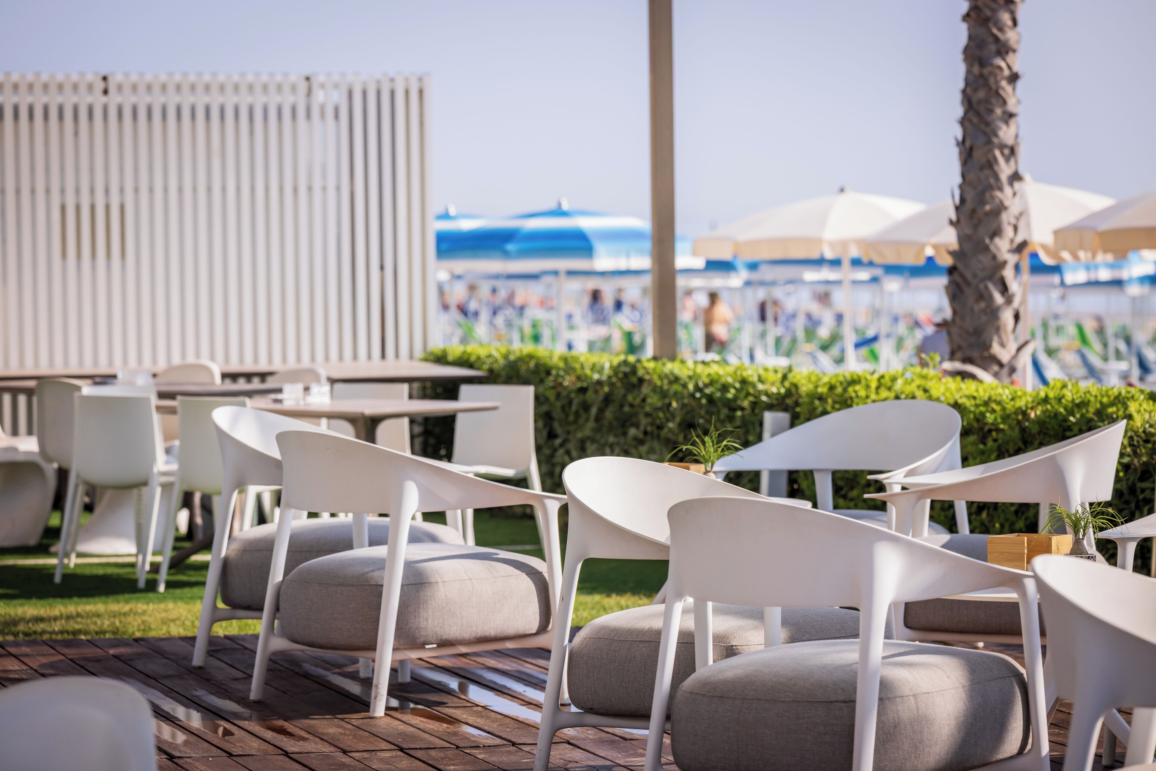 Outdoor seating with white chairs and tables, a wooden deck, and a view of a beach with umbrellas.
