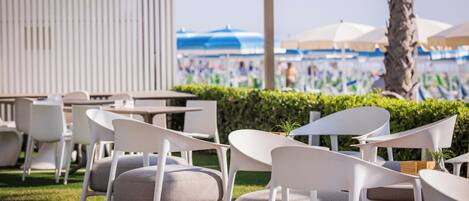 Mobiliario exterior con sillas y mesas blancas, una terraza de madera y vista a una playa con sombrillas.