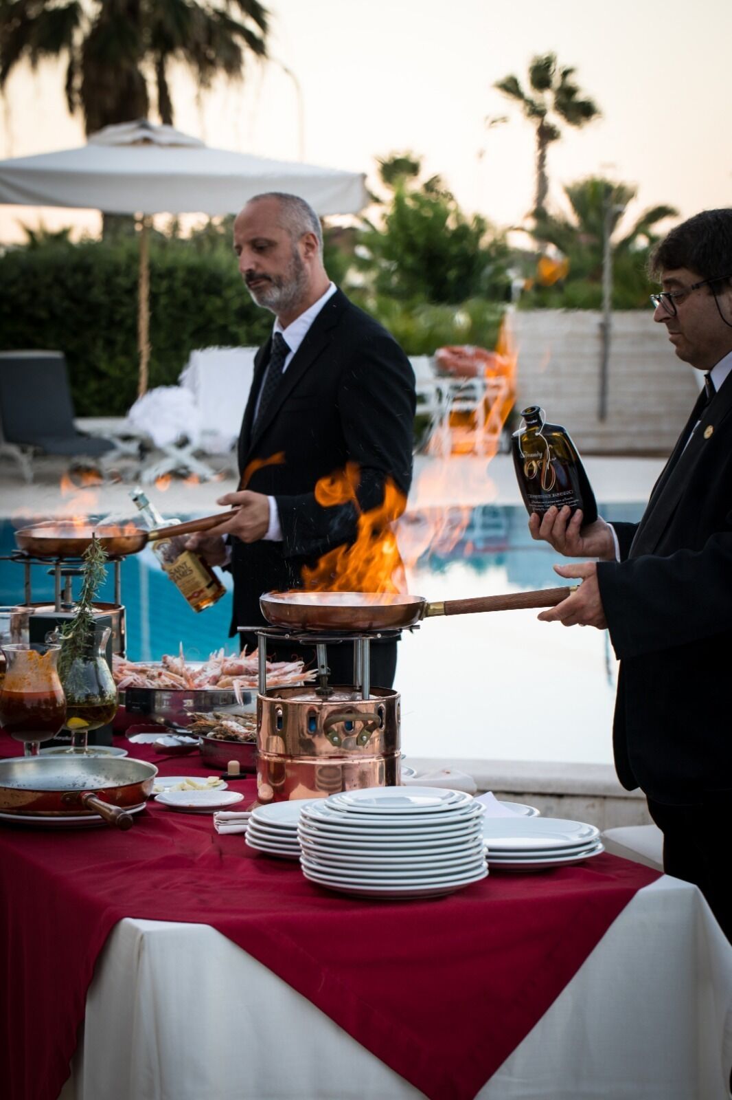 A man grilling food outdoors with a fire, a table with plates and a bottle, and a pool in the background.