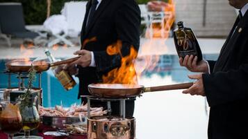 A man grilling food outdoors with a fire, a table with plates and a bottle, and a pool in the background.