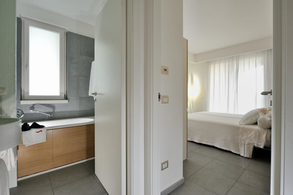 A modern bathroom with a sink, mirror, and towel rack. The room features a bed with white bedding and a window with curtains.