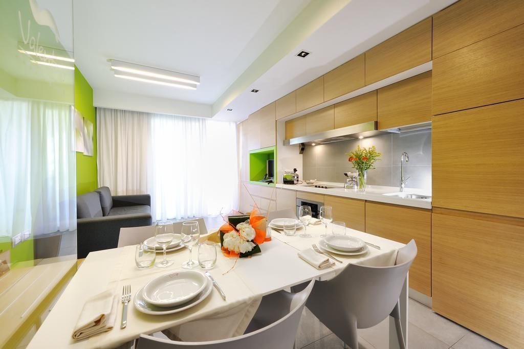 Un comedor moderno con una mesa preparada para cuatro, una cocina al fondo y una pared verde de acento.