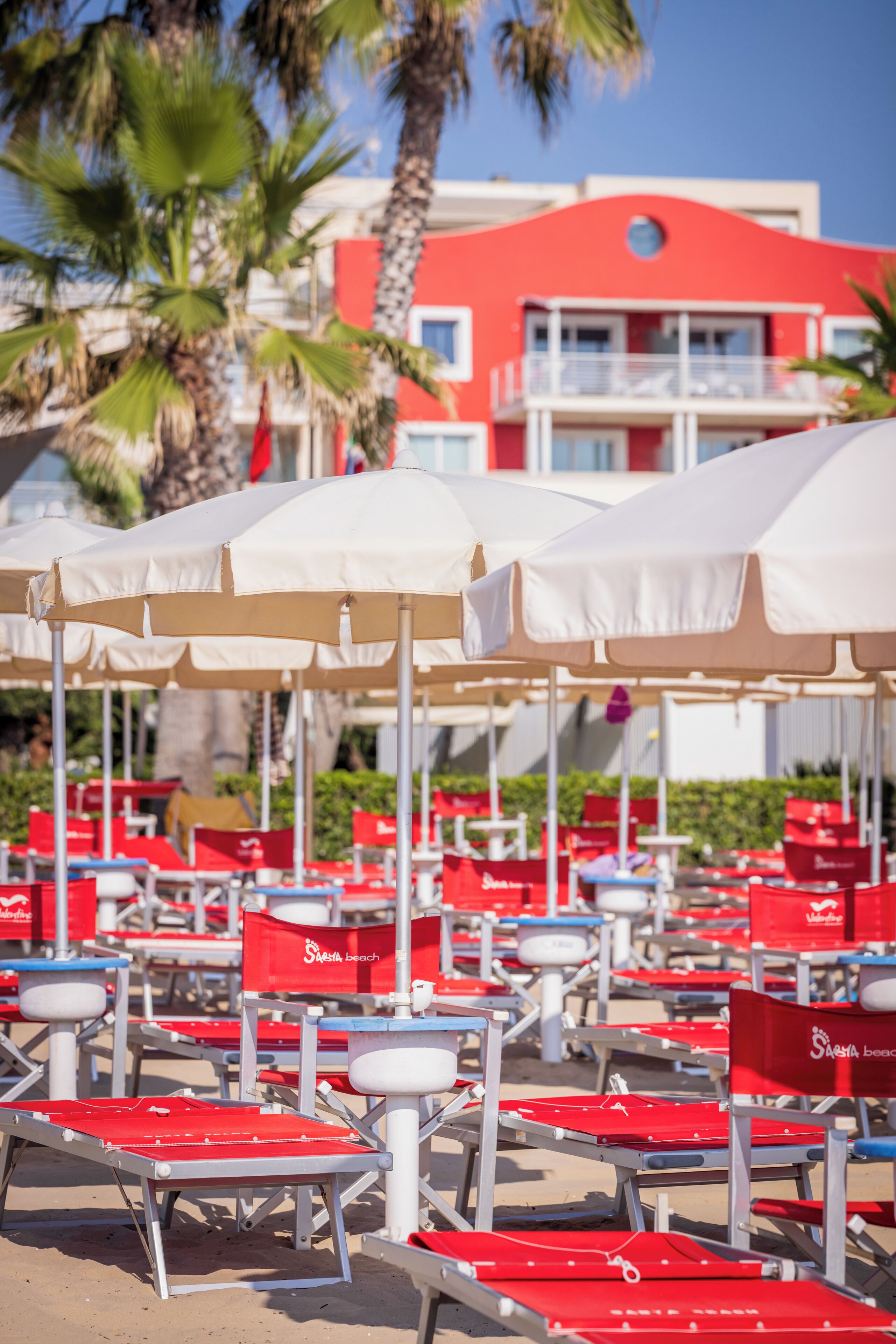 Een strand met rijen rode en witte ligbedden, witte parasols en een rood gebouw op de achtergrond.