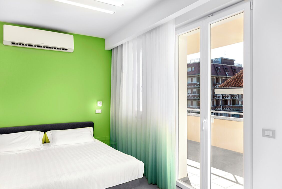 Habitación de hotel con cama, unidad de aire acondicionado, ventana con cortinas y vistas a edificios.