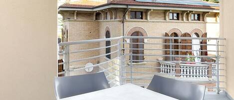 Un balcón con mesa y sillas, vista a un edificio con ventanas arqueadas y barandilla.