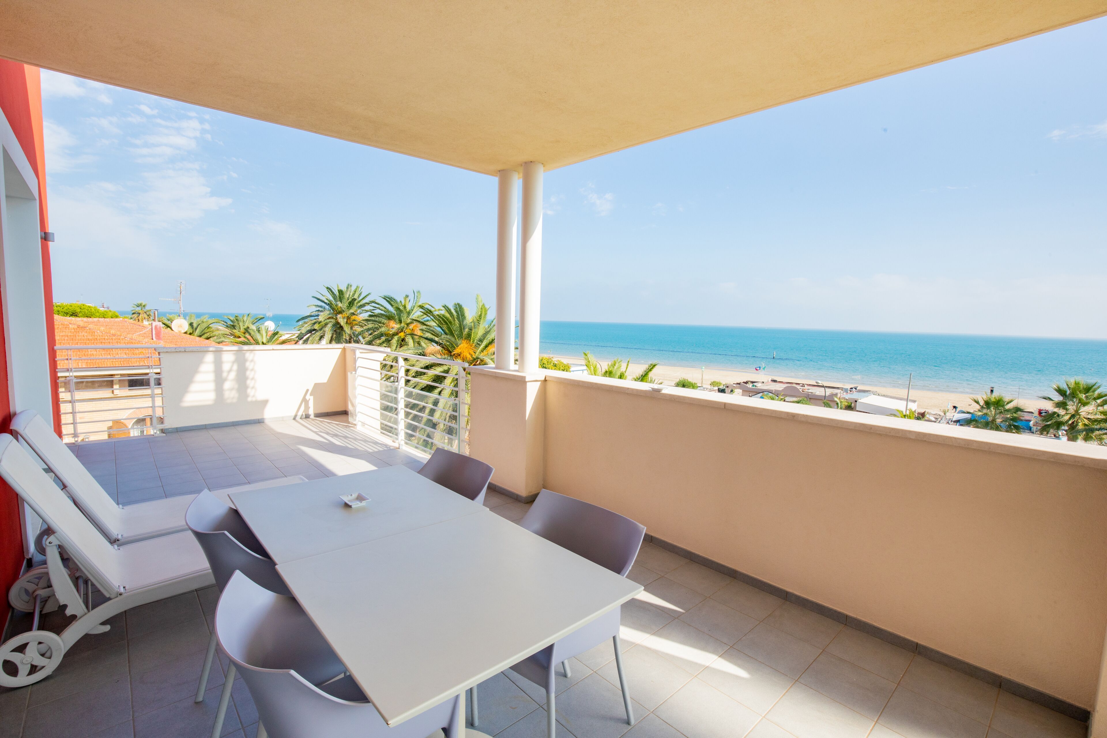 Een balkon met een witte tafel en stoelen, uitkijkend op een strand met palmbomen en de zee.
