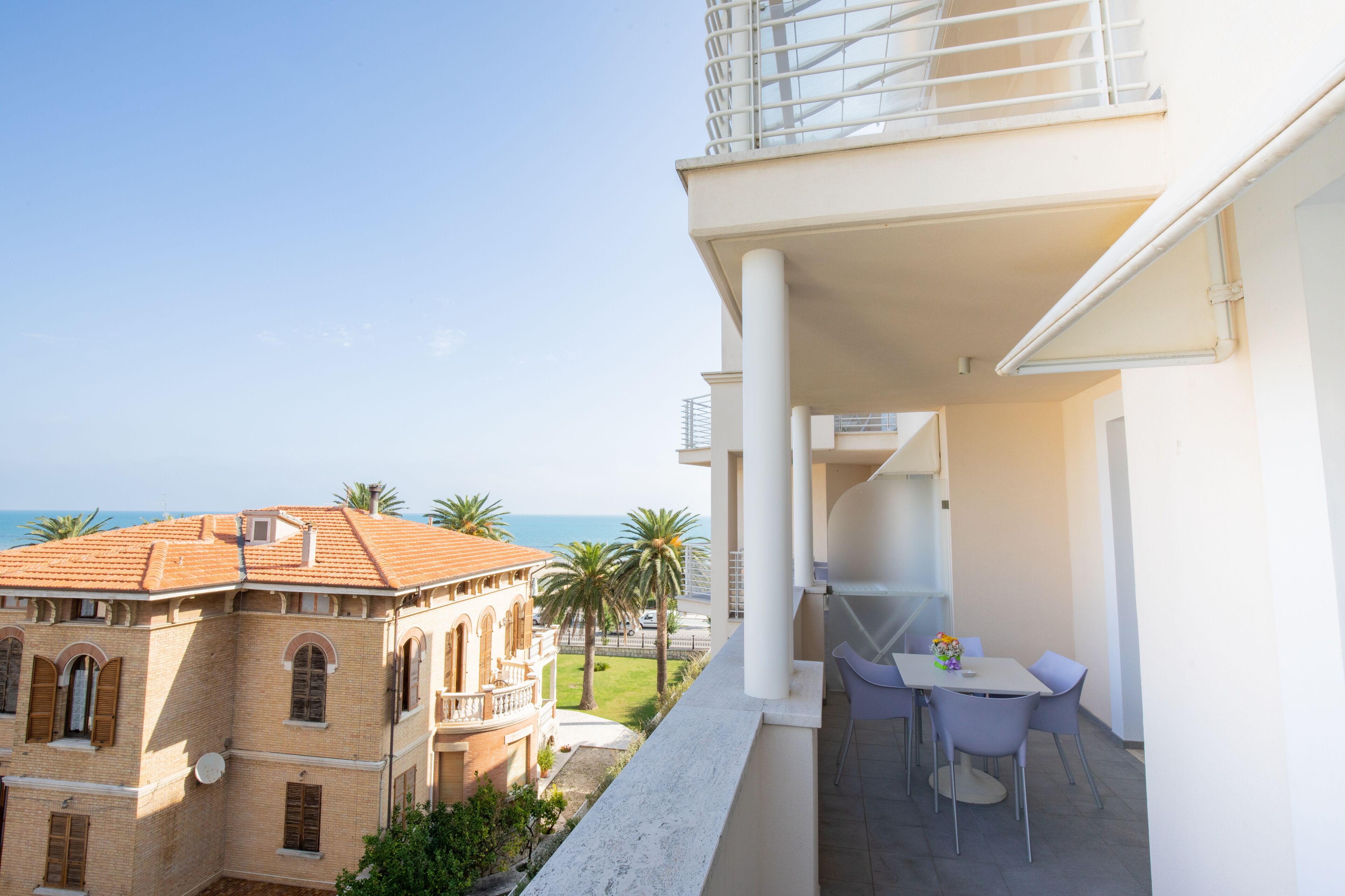 Ein Balkon mit Tisch und Stühlen, mit Blick auf ein Gebäude mit rotem Ziegeldach und Palmen.