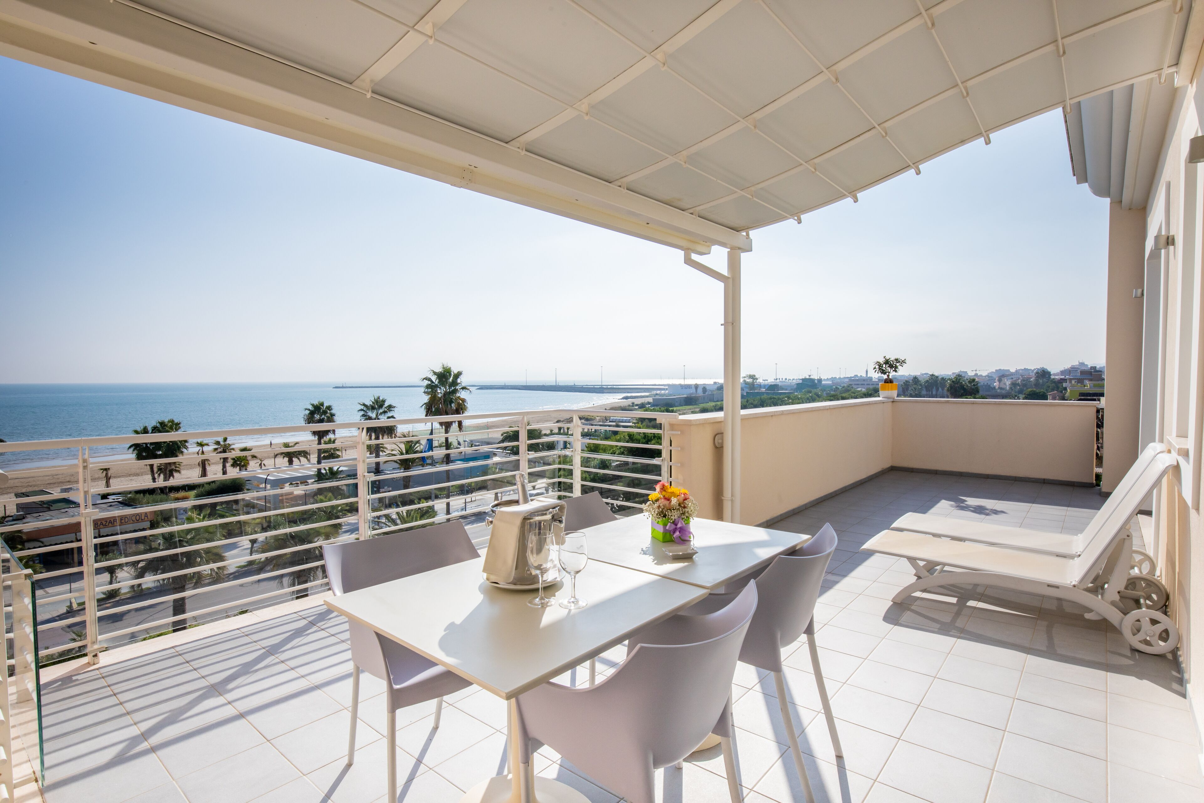 A balcony with a table and chairs, a lounge chair, and a view of the sea and cityscape.