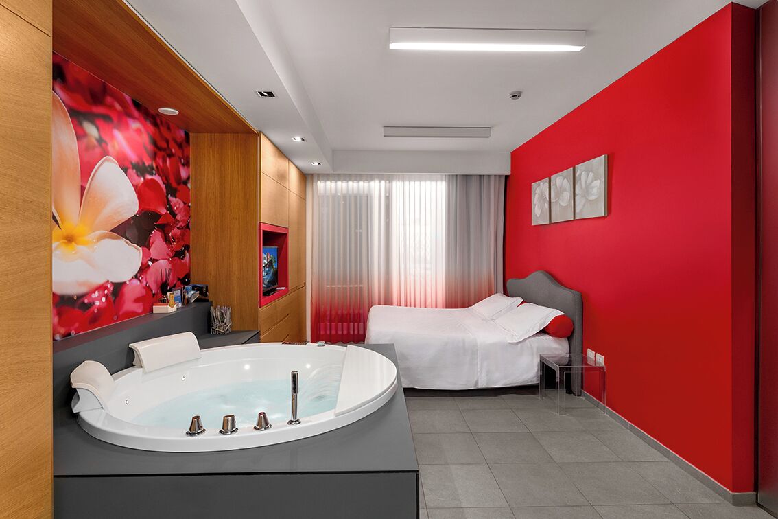 A modern hotel room with a large bathtub, a bed, and a red accent wall.
