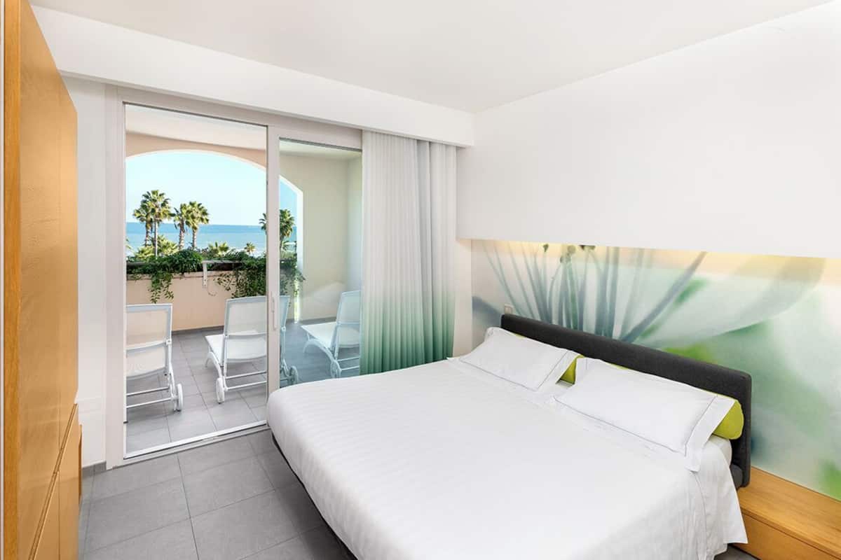 Habitación de hotel con cama, vista al océano, balcón con sillas y puerta corredera.