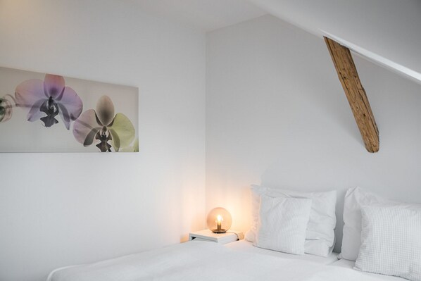 A bedroom with a bed, white pillows, a bedside table with a lamp, and a wall-mounted artwork of flowers.
