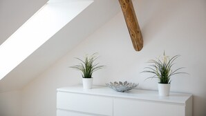 Une commode blanche, ornée de deux plantes en pot et d’un bol décoratif. Une planche de bois est fixée au mur.
