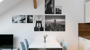 Un espace repas moderne avec une table blanche, des chaises bleues et un vase de fleurs. Au mur, on peut voir des photographies en noir et blanc représentant des ponts et des paysages urbains.