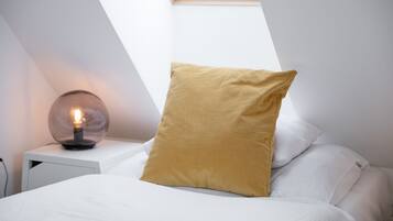 Un dormitorio con tragaluz, lámpara de noche y cama con ropa blanca y almohada color mostaza.
