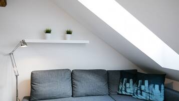 Une chambre de style loft moderne, avec un plafond en pente, un canapé gris, une lampe sur pied et deux plantes en pot posées sur une étagère blanche.