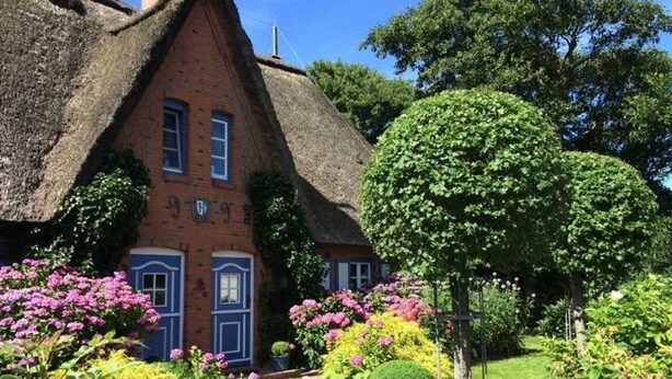 Een huis met een rieten dak en gevel van baksteen, omgeven door goed onderhouden tuinen en diverse bloeiende planten.