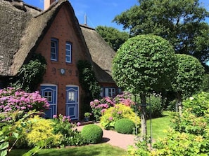 A traditional thatched-roof house with a brick facade, surrounded by well-maintained gardens and a variety of flowering plants. - House half 41 Against the wind (ID 162) (Sankt Peter-Ording)