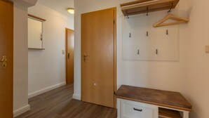 Un pasillo con suelo de madera, una puerta de madera, un perchero con ganchos y un banco con cajones.