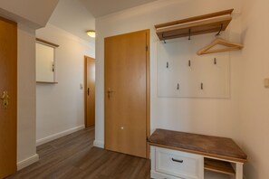 A hallway with wooden flooring, a wooden door, a coat rack with hangers, and a bench with drawers.