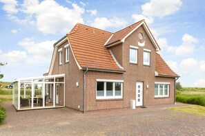 Une maison à deux étages, avec une façade en briques, un toit en tuiles et une véranda en verre aménagée avec du mobilier d’extérieur.