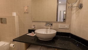 A modern bathroom with a white sink, black countertop, and a wooden shelf with towels. - The Solitaire Express (Dehradun)