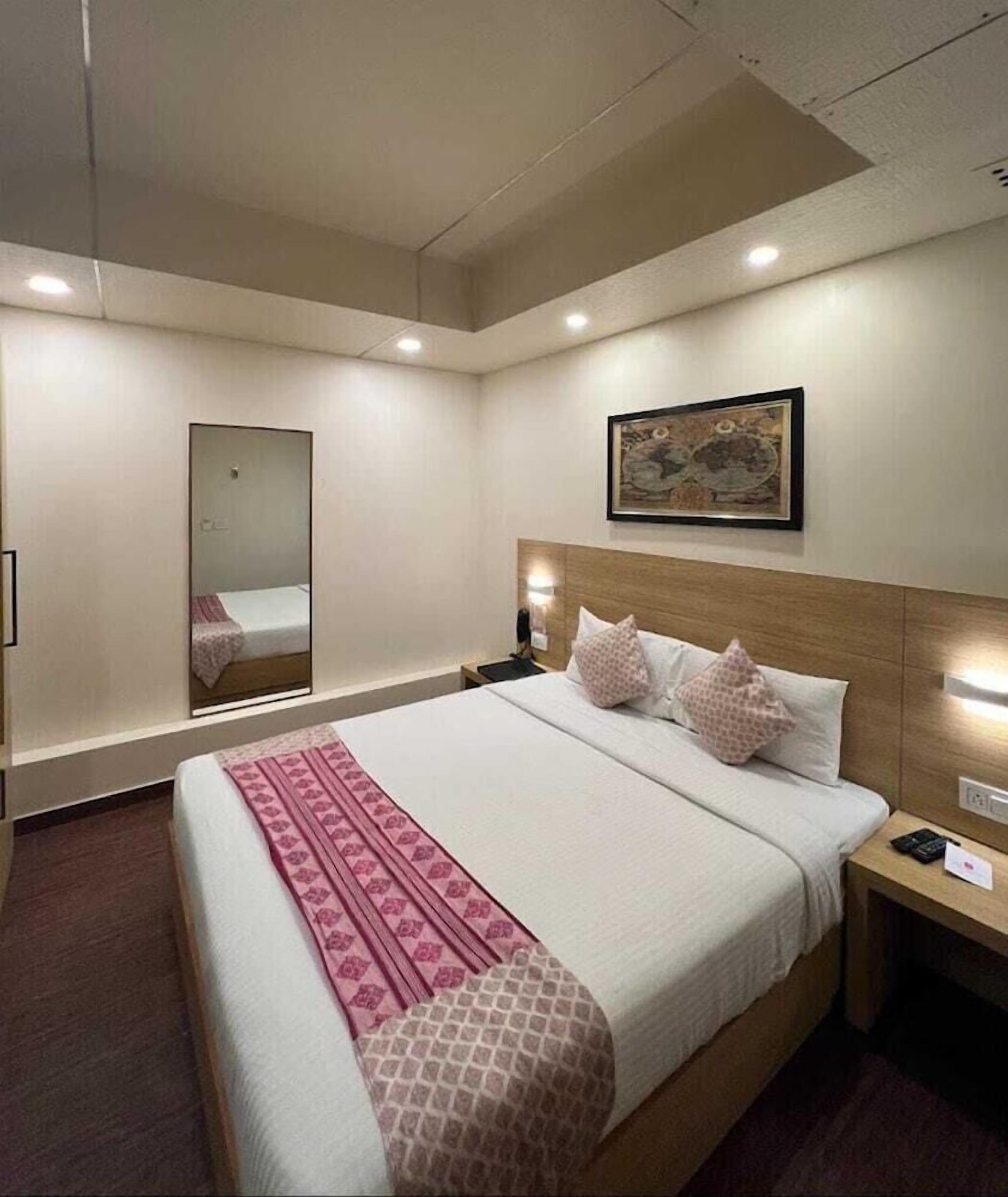 A hotel room with a bed, a nightstand, a wall-mounted lamp, a mirror, and a framed picture.