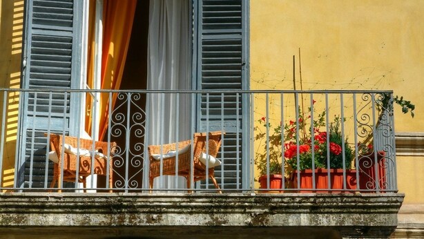 Un balcon doté d’une balustrade en fer forgé, une petite table avec une chaise, et des plantes en pot ornées de fleurs rouges.