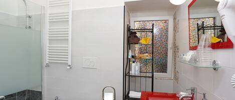 A bathroom with a glass shower enclosure, a bidet, a toilet, a red sink, and a glass shelf with toiletries.