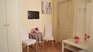 Une petite pièce avec deux chaises blanches, une table ornée d’un bouquet de fleurs roses, un téléviseur accroché au mur et une porte dotée d’un miroir.