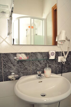 A bathroom with a white sink, a glass shelf with toiletries, a hairdryer, and a shower area. - Locanda al Viminale (Rome)