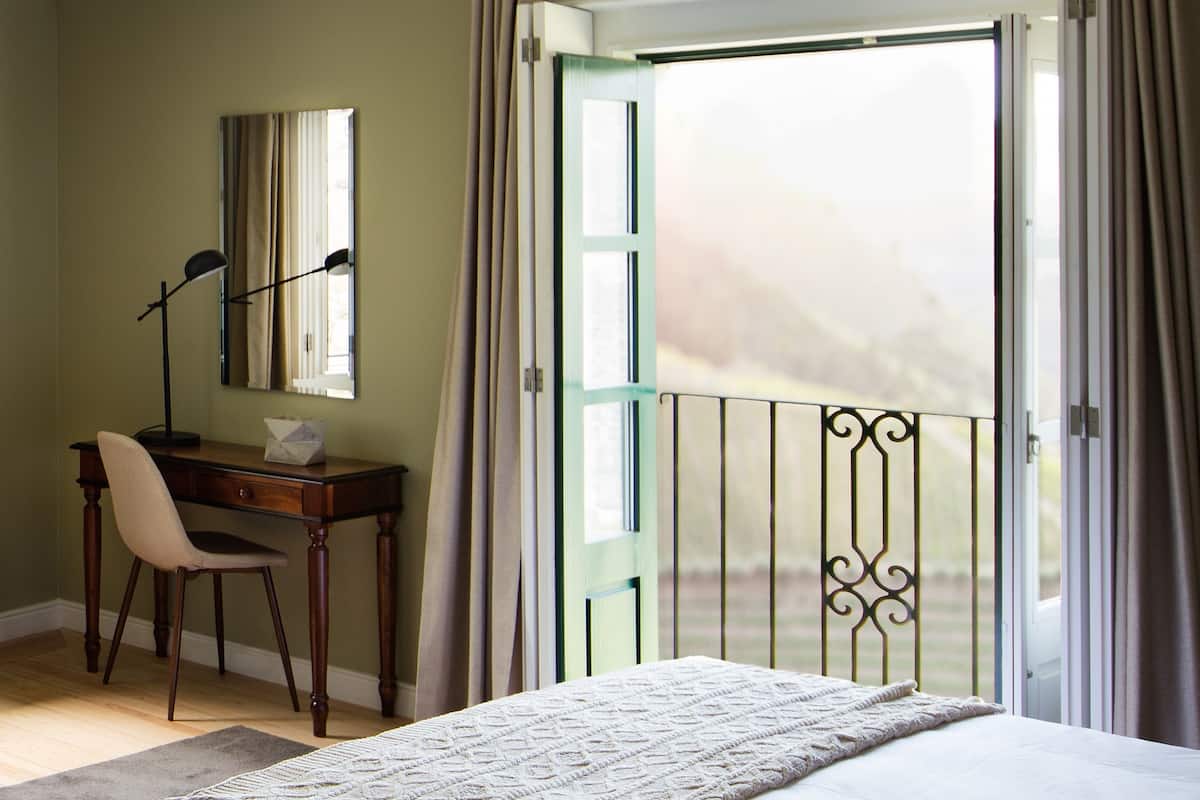 Ein Schlafzimmer mit einem Bett, einem Schreibtisch mit Stuhl, einem Fenster mit Vorhängen und einem Balkon mit Geländer.