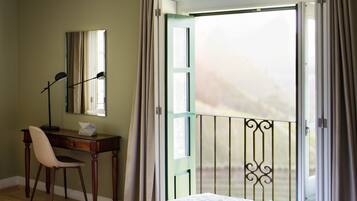 Quarto com cama, escrivaninha com cadeira, janela com cortinas e varanda com guarda-corpo.