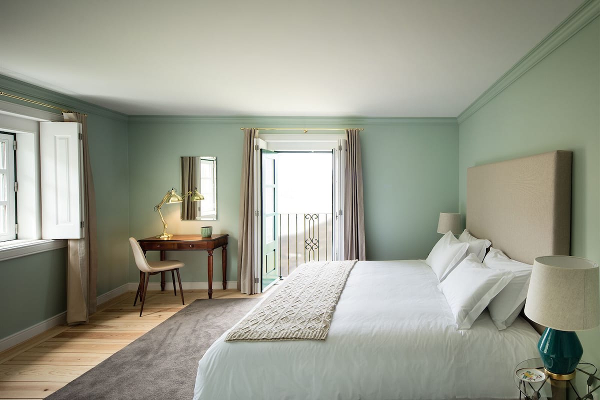 Ein Schlafzimmer mit Bett, Nachttisch, Lampe, Stuhl, Schreibtisch und einem Fenster mit Vorhängen.