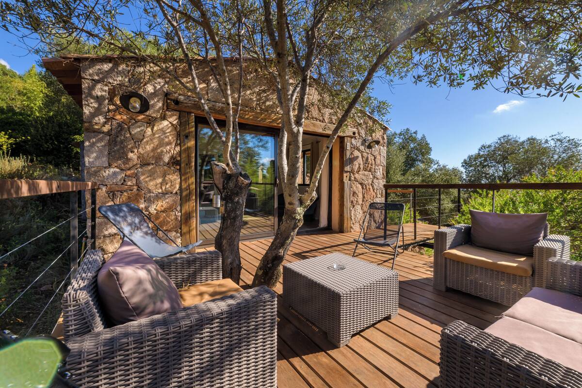 A wooden deck with wicker furniture, a stone building with a glass door, and a tree in the background.