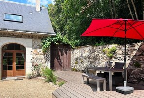 Ein Steinhhaus mit einer Holzterrasse, einem roten Sonnenschirm und einer Bank.