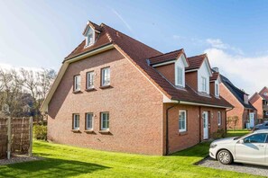 Une maison à deux étages, construite en briques, avec un toit en pente, de nombreuses fenêtres et une voiture garée dans l’allée.