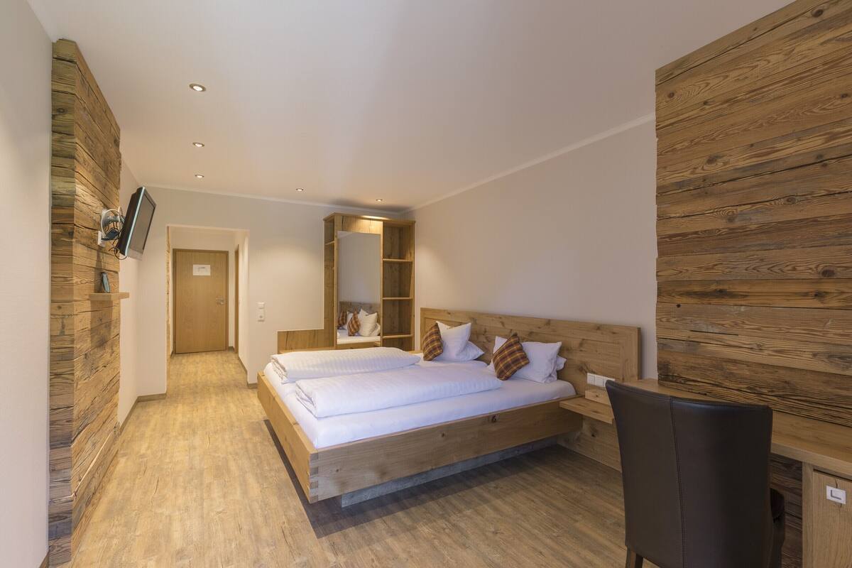 A double bed with a wooden headboard, a bedside table, and a chair. There is a TV mounted on the wall and a wooden panel on the right side.