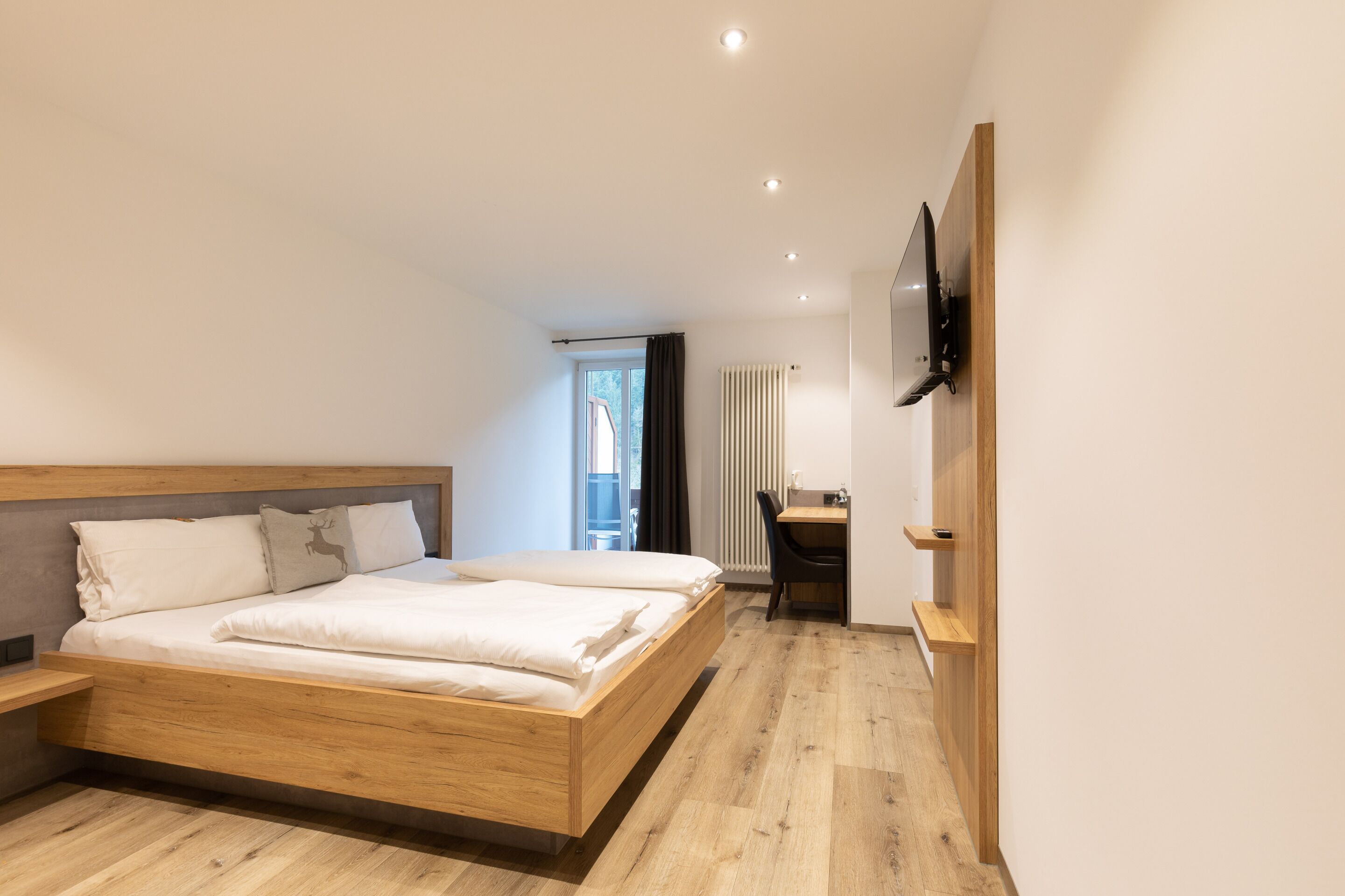 A modern bedroom with a wooden bed, a desk, and a TV mounted on the wall.