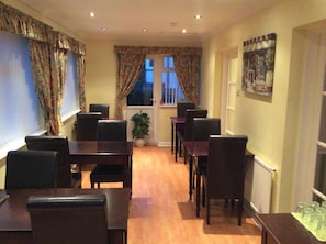 A dining area with wooden tables and chairs, a potted plant, and a window with curtains. - White Lodge Guest House (Skegness)