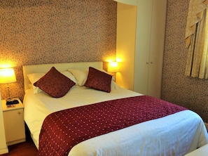 A hotel room with a bed, two bedside lamps, a nightstand, and a patterned wall. - White Lodge Guest House (Skegness)