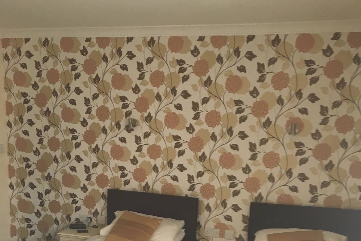 Two single beds with white linens and brown pillows, a nightstand between them, and a floral-patterned wallpaper on the wall.