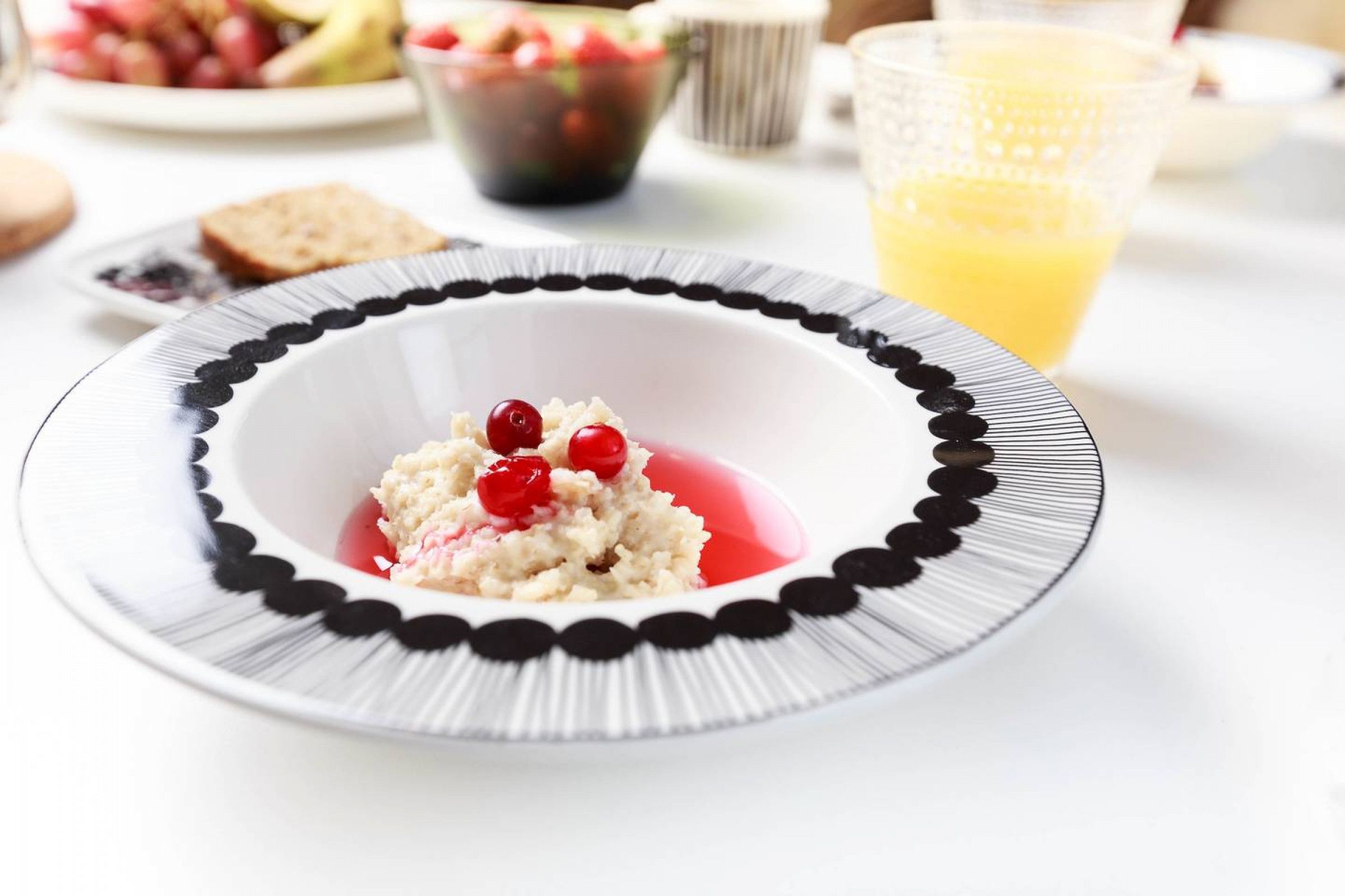 A white plate with a black rim containing a dessert with red berries, a glass of orange juice, and a bowl of mixed berries in the background.