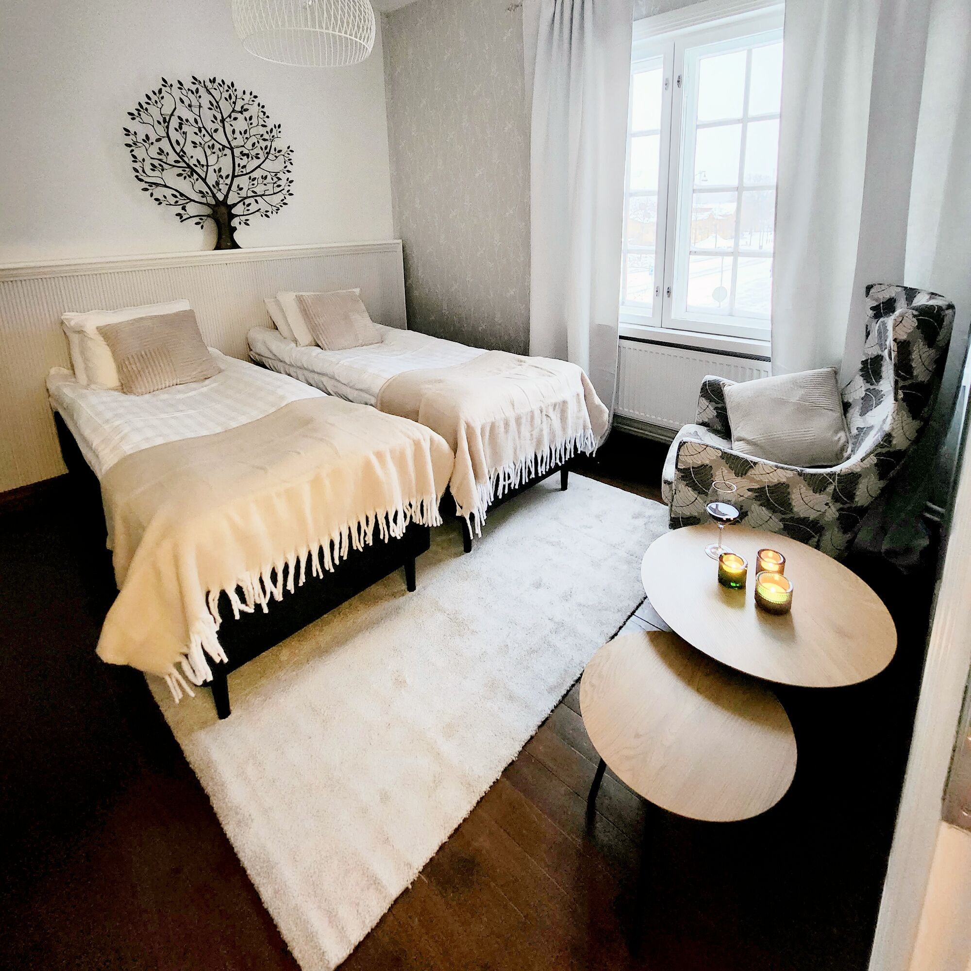 Een slaapkamer met twee eenpersoonsbedden, een stoel, een rond tafeltje met kaarsen en een wanddecoratie van een boom.