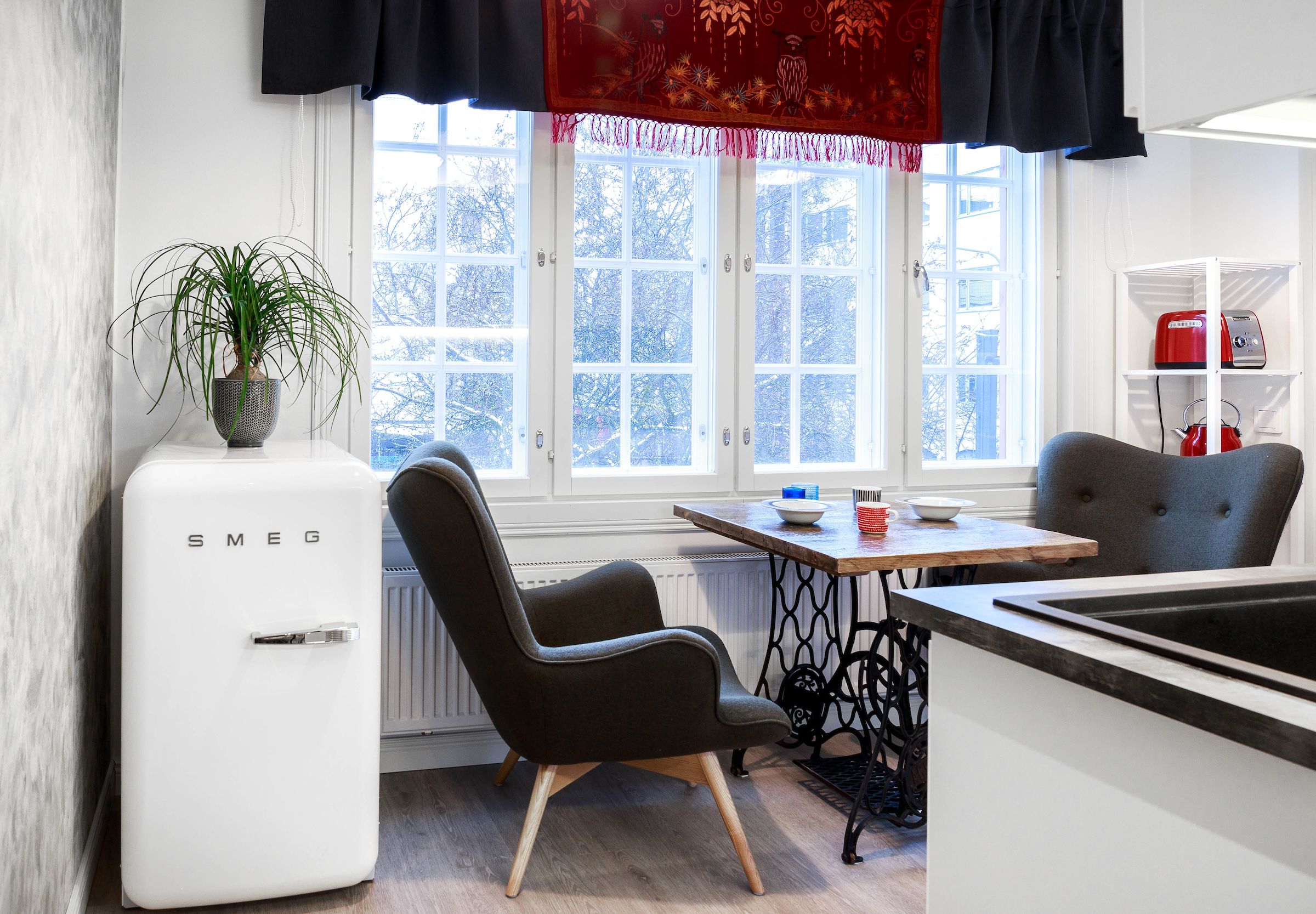 Una cocina con un refrigerador blanco, una mesa de comedor, una silla negra y una ventana con vistas a árboles.