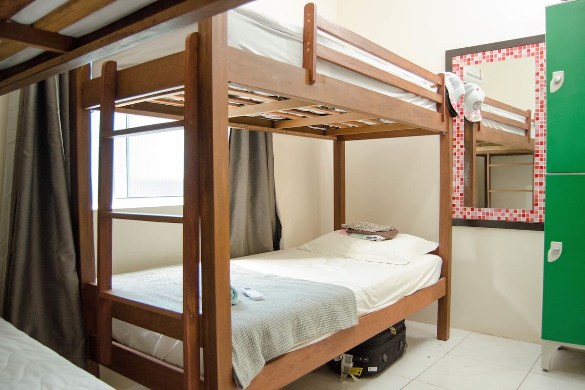 Una cama litera con estructura de madera, una cama individual abajo y un armario verde con salpicadero de azulejos.