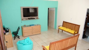 A living room with a TV, wooden furniture, a teal bean bag, and a wooden bench with yellow cushions.