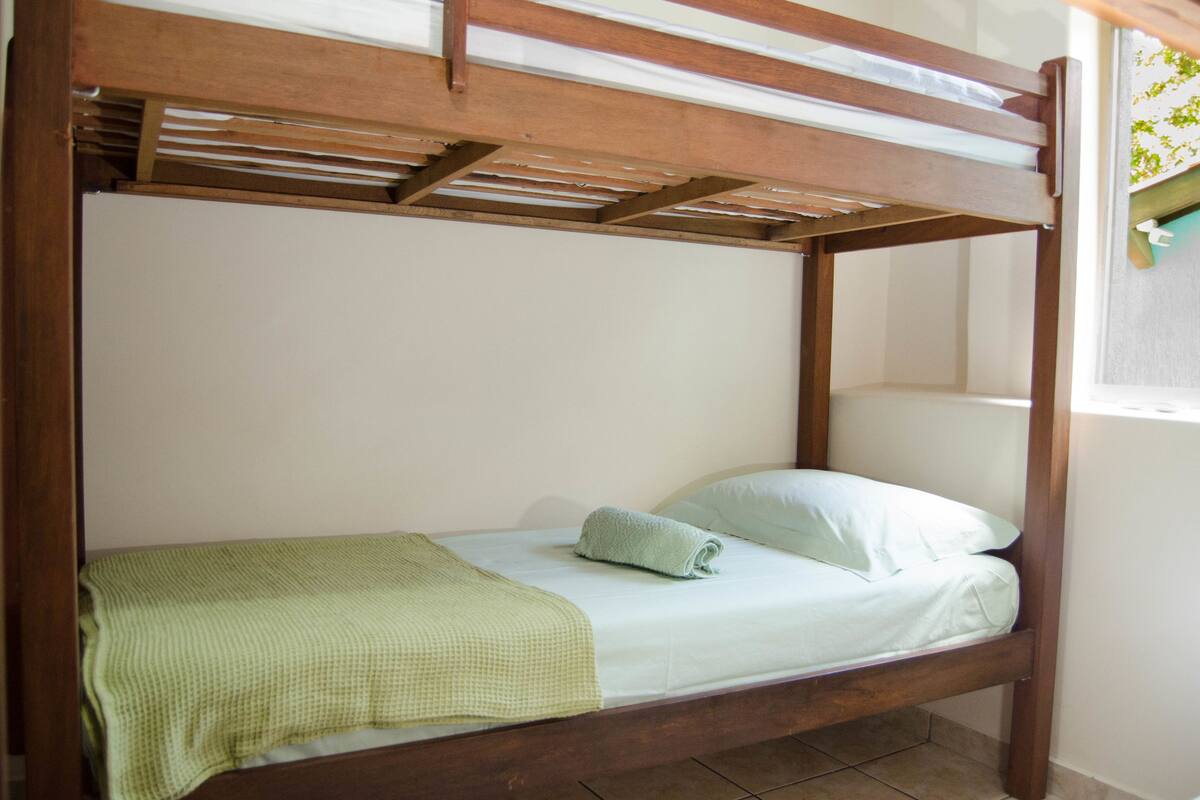 Una cama individual con estructura de madera, una manta verde y una almohada blanca en una habitación con ventana.