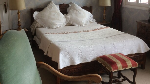 Un dormitorio con una cama, dos lámparas de noche, una silla, un banco y una ventana con cortinas.