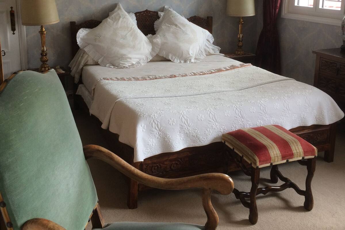 A bedroom with a bed, two bedside lamps, a chair, a bench, and a window with curtains.